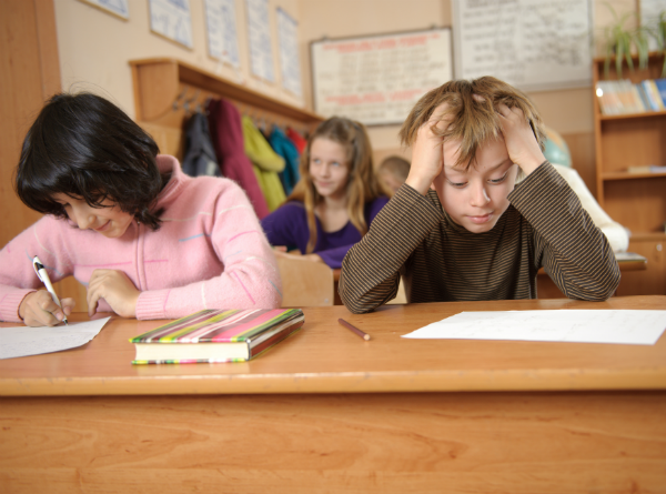 student uvádí u svého stolu, sevře hlavu rukama a dívá se dolů na prázdný list papíru.