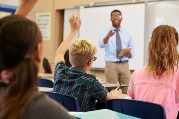 um professor aponta para um aluno que está levantando a mão na aula para responder a uma pergunta.