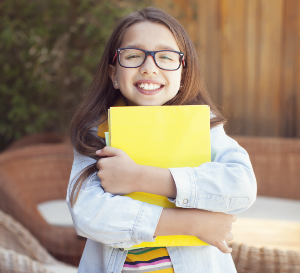 A female elementary student stands clutching a yellow folder to her chest and stomach.