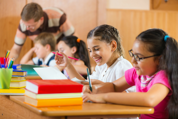 A row of elementary students sit at a desk, using pen and paper to work on project based learning examples.