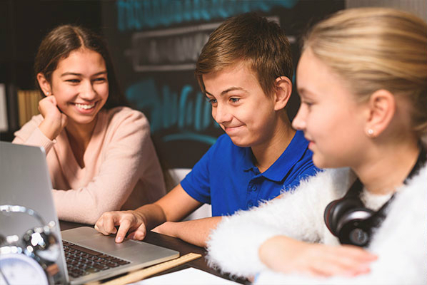 Three elementary students crowd around a silver Mac laptop, completing a project based learning project together.