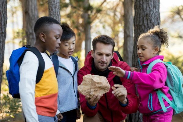 Three young elementary students gather around their teacher outside, who's showing and talking about a kind of rock.