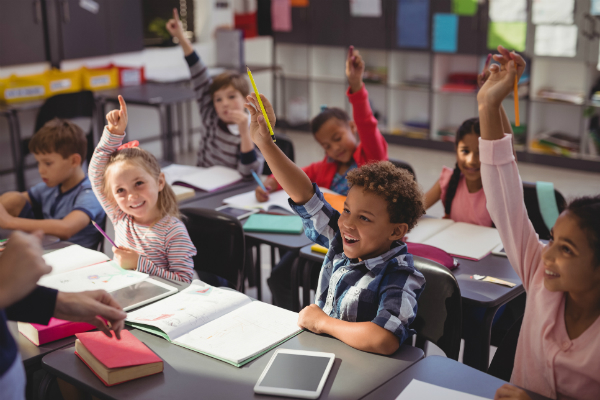 Students sit in a classroom, with raised hands, ready to answer the teacher's questions