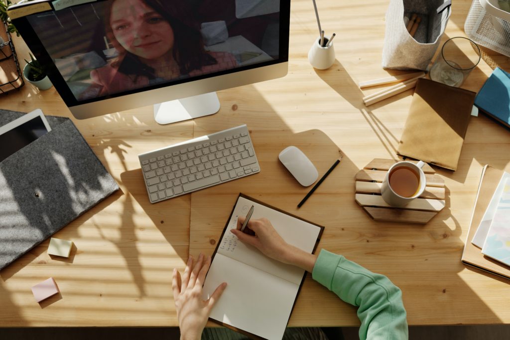 Two teachers chatting over a video call while one writes in a notebook on her desk.